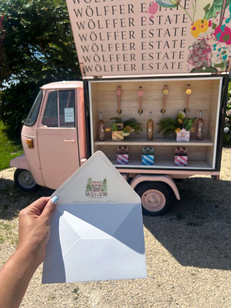 Luxury wedding invitation envelope with custom illustrated liner held in front of Wölffer Estate rosé truck in the Hamptons