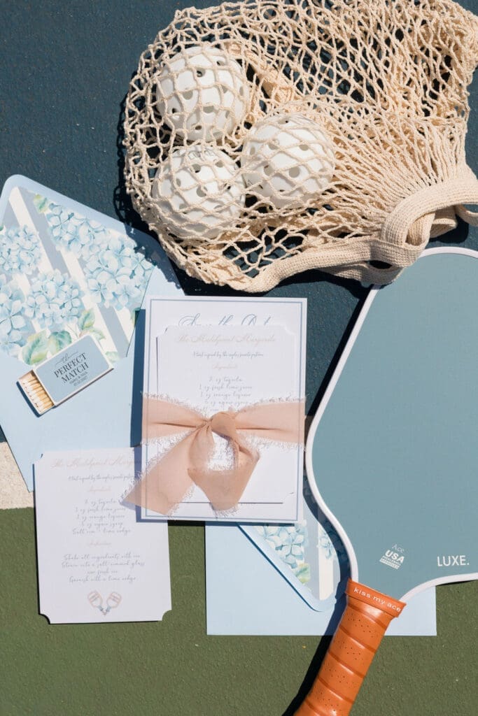 Luxury wedding stationery featuring blue watercolor hydrangea envelope liners and custom matchboxes styled on a pickleball court.