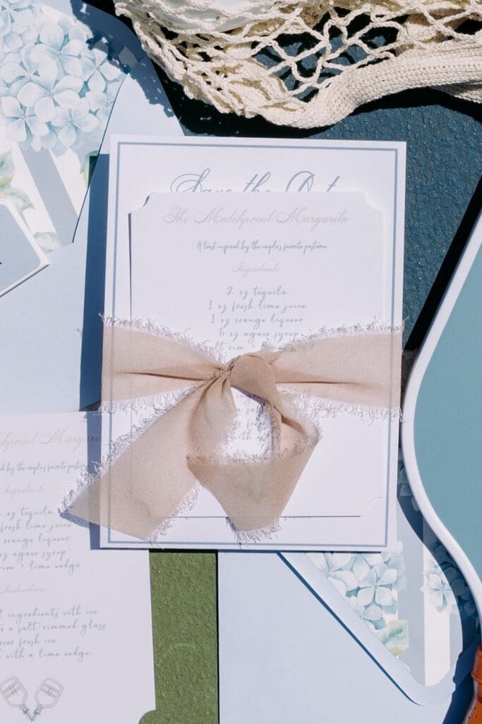 Closeup of a luxury save the date suite featuring a custom cocktail recipe card tied with soft neutral ribbon.