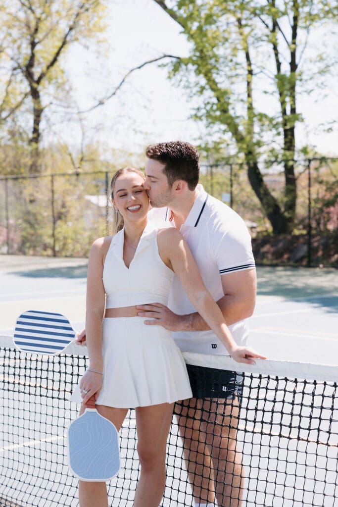 Couple posing at a pickleball net during a light and airy engagement photoshoot with blue paddles.