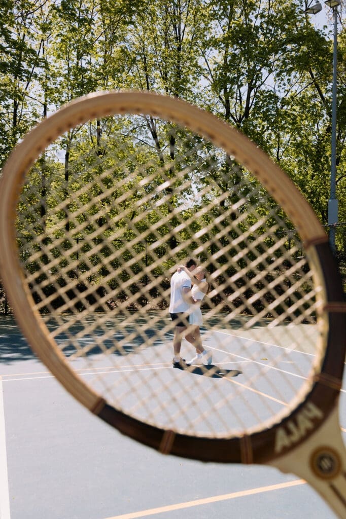 Couple embracing on a pickleball court framed through a vintage wooden racket during an editorial engagement photoshoot.