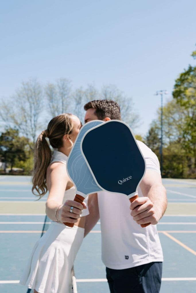 Couple holding blue pickleball paddles during a playful engagement photoshoot on an outdoor court.