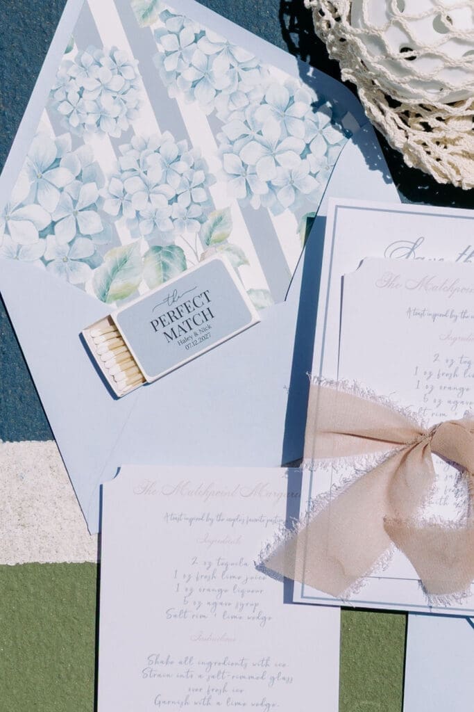 Luxury wedding stationery featuring blue watercolor hydrangea envelope liners and custom matchboxes styled on a pickleball court.
